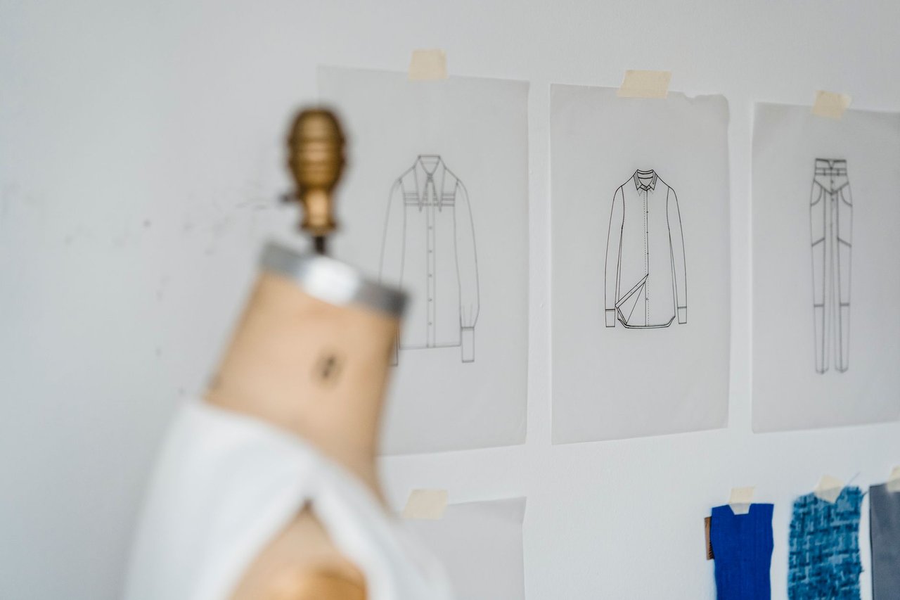 Manequim desfocado em um ateliê iluminado, cercado por esboços de roupas e amostras coloridas de tecidos.